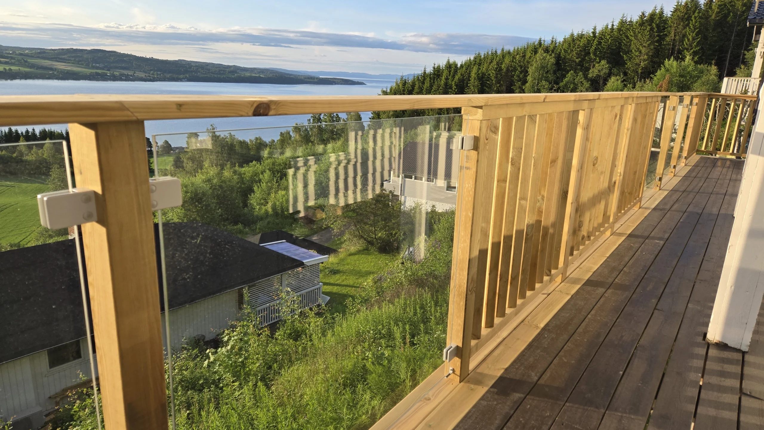 Altanrække med glas og ribber langs en smal altan med udsigt over skov, vand og kuperet landskab.