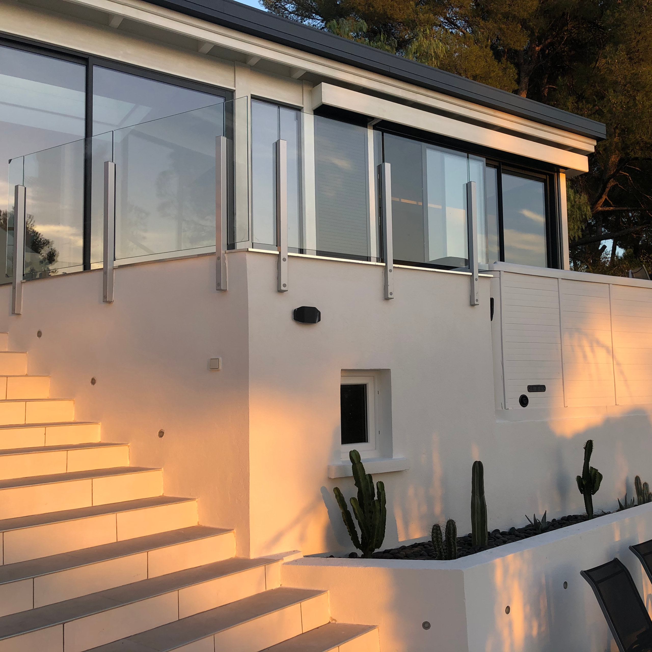 Yttertrappa vid ett vitt hus med glas- och aluminiumräcke där trappstegens höjd kan behöva kontrolleras efter vintern