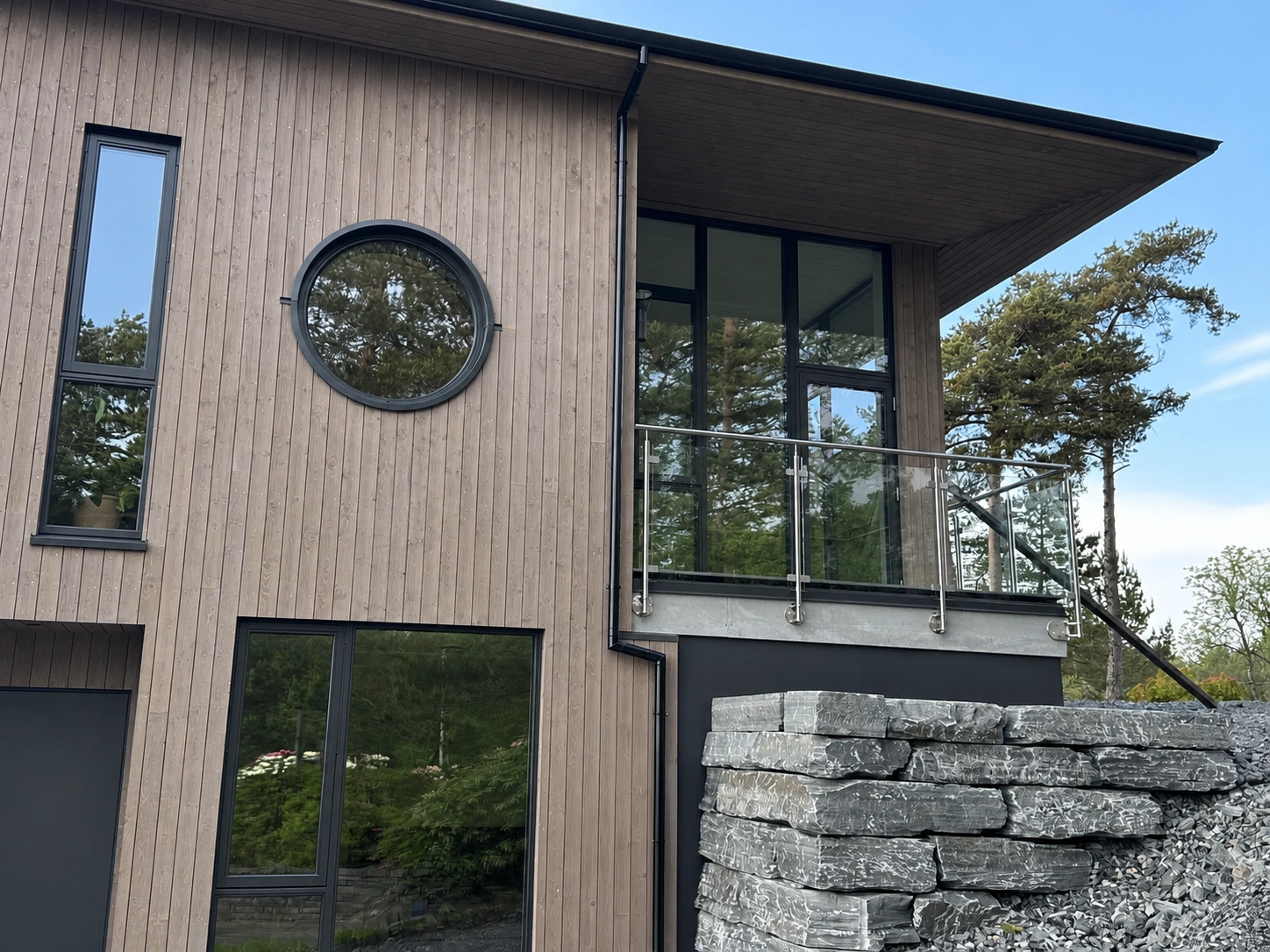 Modernt trähus med glasräcke på balkong, svarta fönsterpartier och naturstensmur framför fasaden.