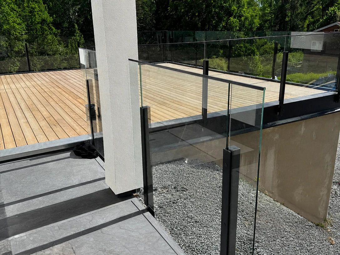 Hörnparti på terrass med glasräcke i svart aluminium, grått klinkergolv och trätrall i anslutning.
