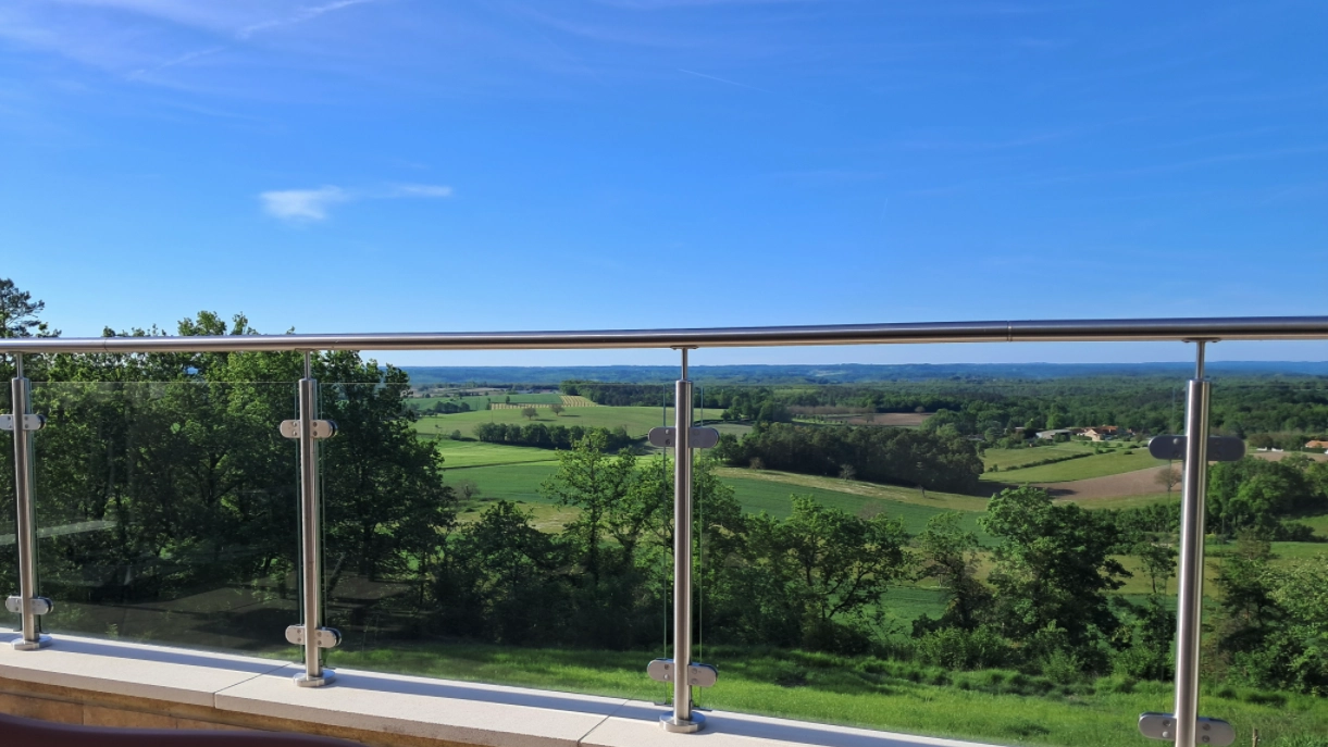Glasgelænder med rustfrie stolper på en terrasse med udsigt over et åbent landskab, monteret som faldsikring langs kanten.