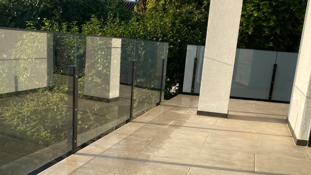 Glasgelænder med tonet sikkerhedsglas på en terrasse ved en hvid facade.