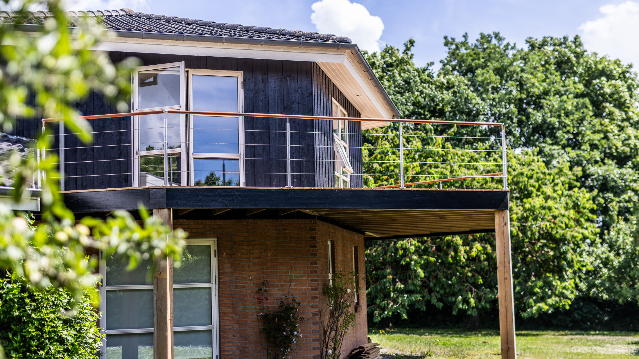 Villa sombre avec une terrasse surélevée et une balustrade en câble métallique avec des traverses en bois, vue depuis le jardin.