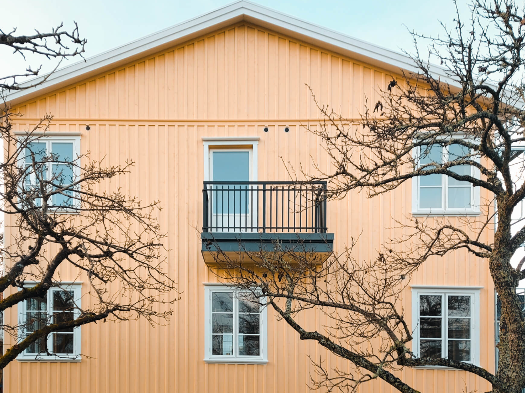 Petit balcon avec une balustrade classique en aluminium forgé, sur une ancienne façade en bois jaune.