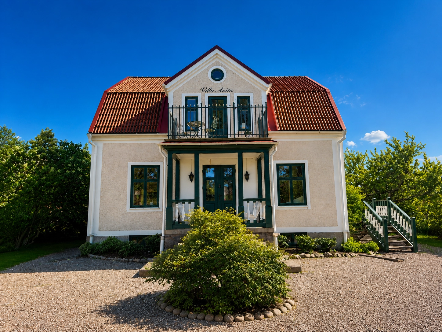 Ljus villa med rött tegeltak, mörkgröna detaljer och smidesinspirerat balkongräcke ovanför entrén.