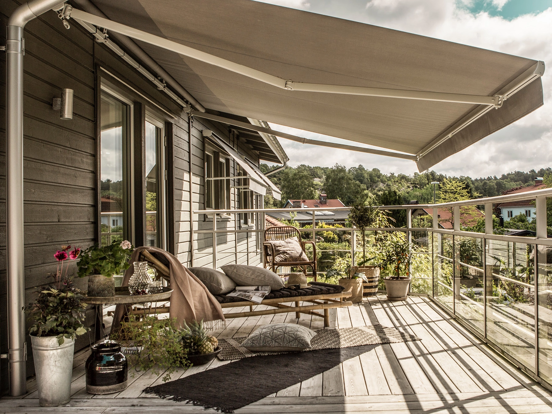 Terrassmarkis över träaltan med glasräcke, soffhörna, krukväxter och mörk träfasad längs huset.