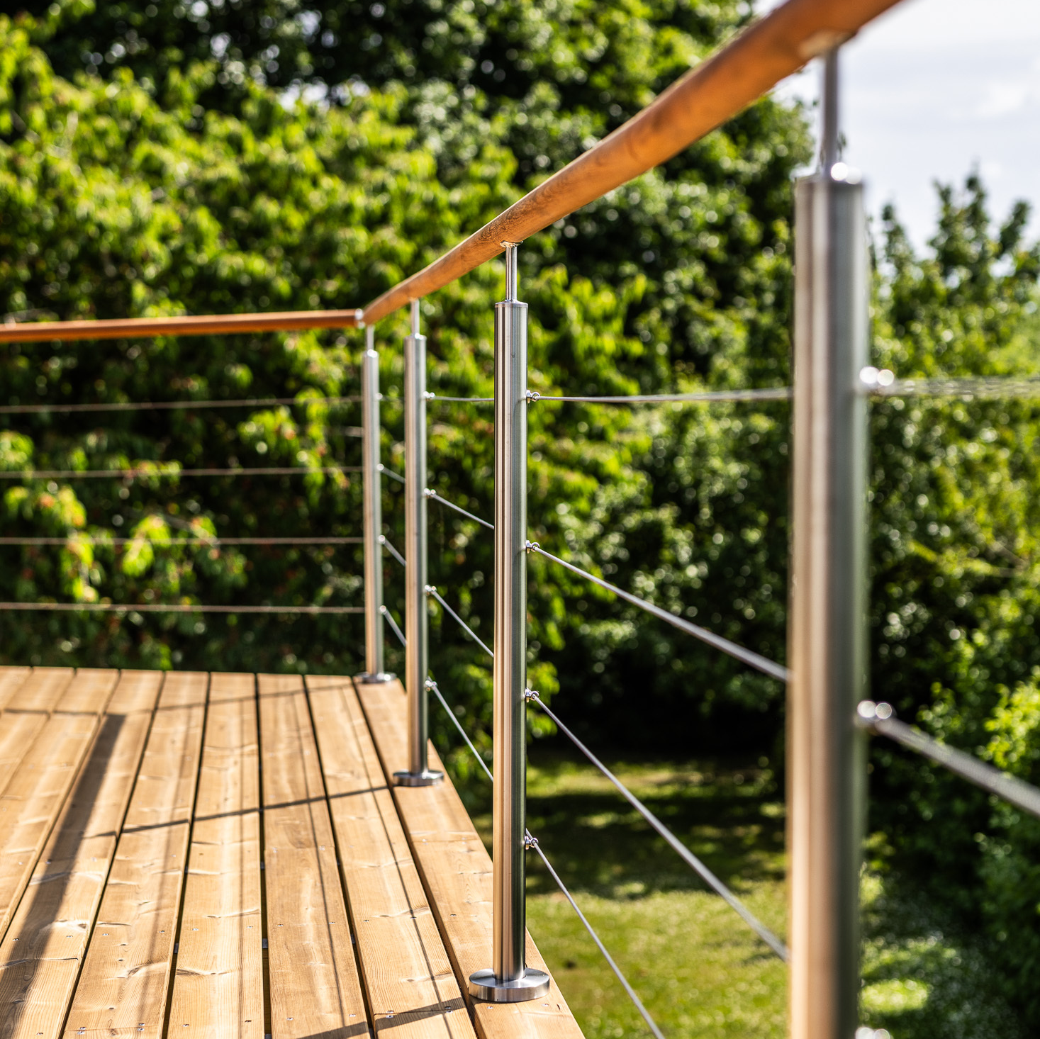 Des poteaux en acier inoxydable avec des câbles métalliques et des traverses en bois le long de la terrasse en bois, avec des arbres fruitiers et de la verdure en arrière-plan.