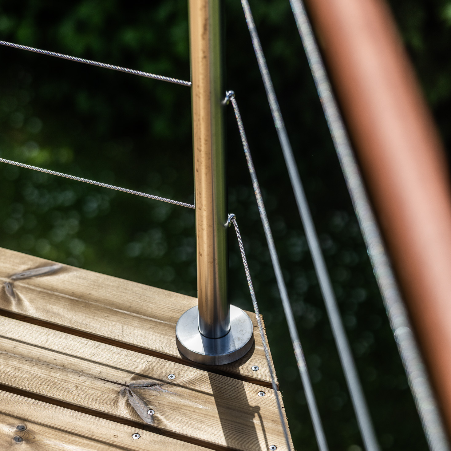 Gros plan sur un poteau en inox muni d'une base, installé au-dessus d'une terrasse en bois, avec des câbles et une traverse en bois.