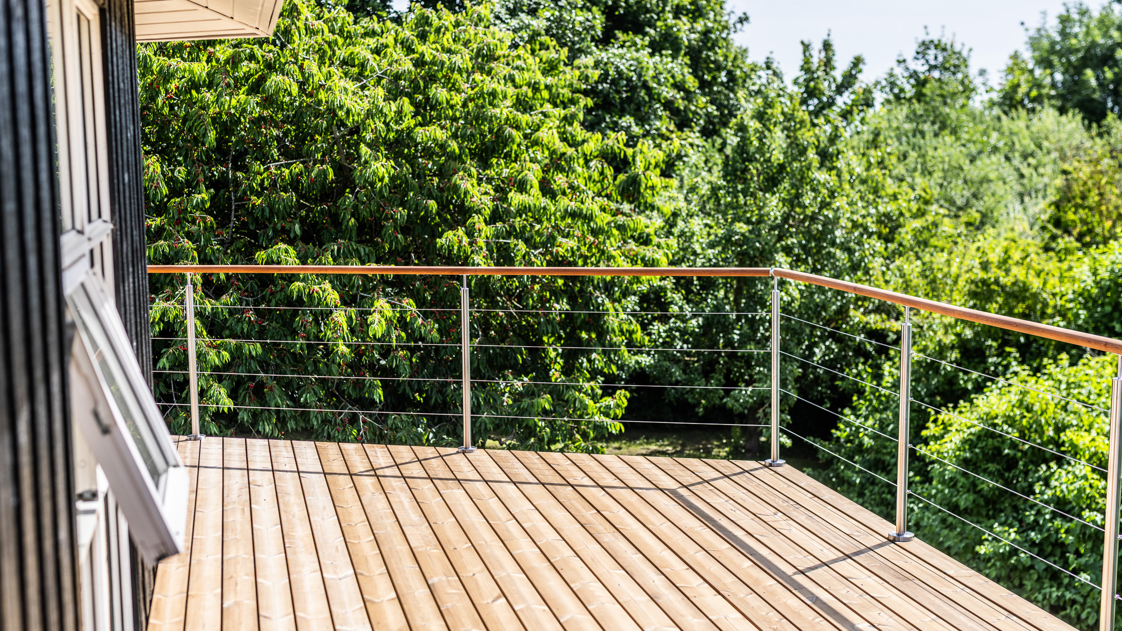 Une balustrade en câble métallique avec des poteaux en acier inoxydable et des traverses en bois, entourant une terrasse surélevée en bois offrant une vue sur un jardin verdoyant.