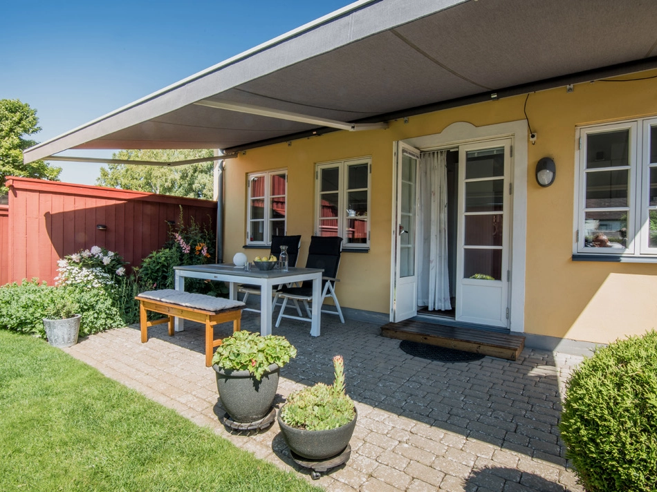 Grå terrassmarkis utfälld över stenlagd uteplats vid gult hus med bord, bänk, blomkrukor och gräsmatta.