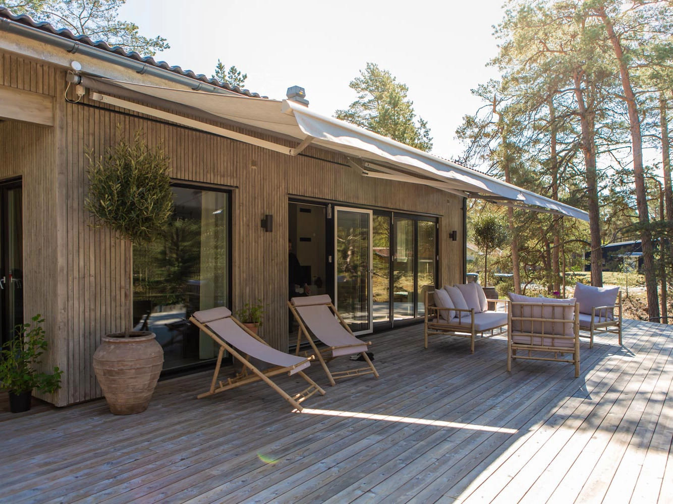 Stor terrassmarkis över träaltan vid träklätt hus med solstolar, loungemöbler, krukor och tallar runt uteplatsen.