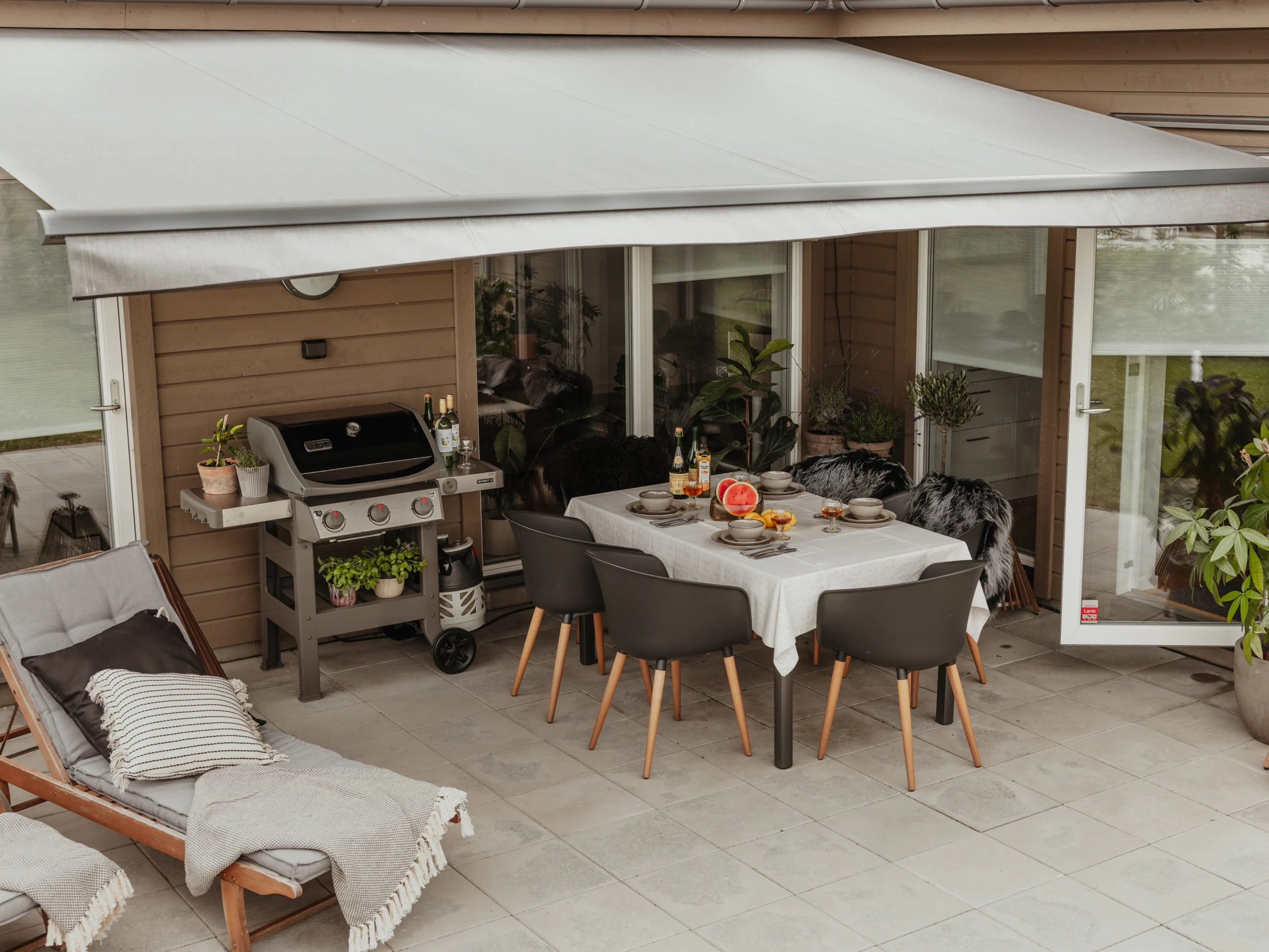 Utfälld terrassmarkis över stenlagd uteplats med matbord, grill, växter och glasdörrar mot träfasad.
